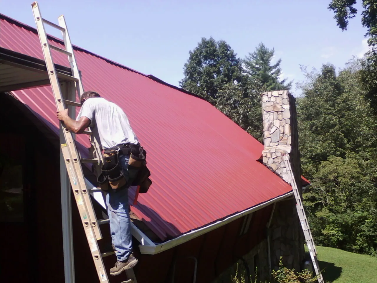 Roofer on a ladder beside a red metal roof and stone chimney — Soffit & Fascia Repair by Roofing Brothers in Eddyville